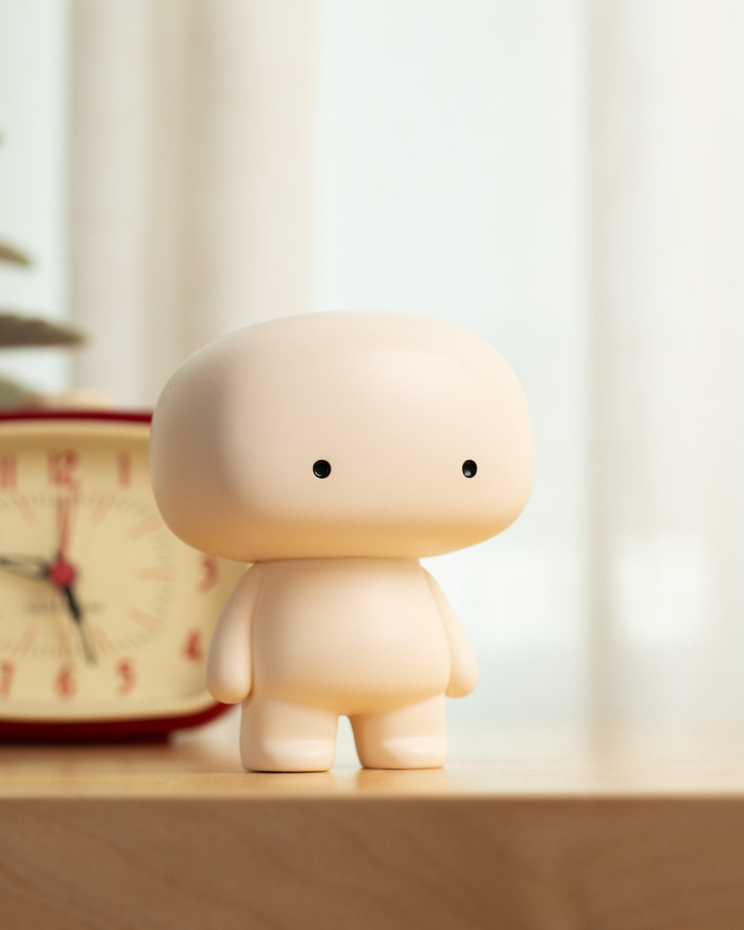 Blumo figure on a wooden shelf beside a vintage alarm clock and potted plant